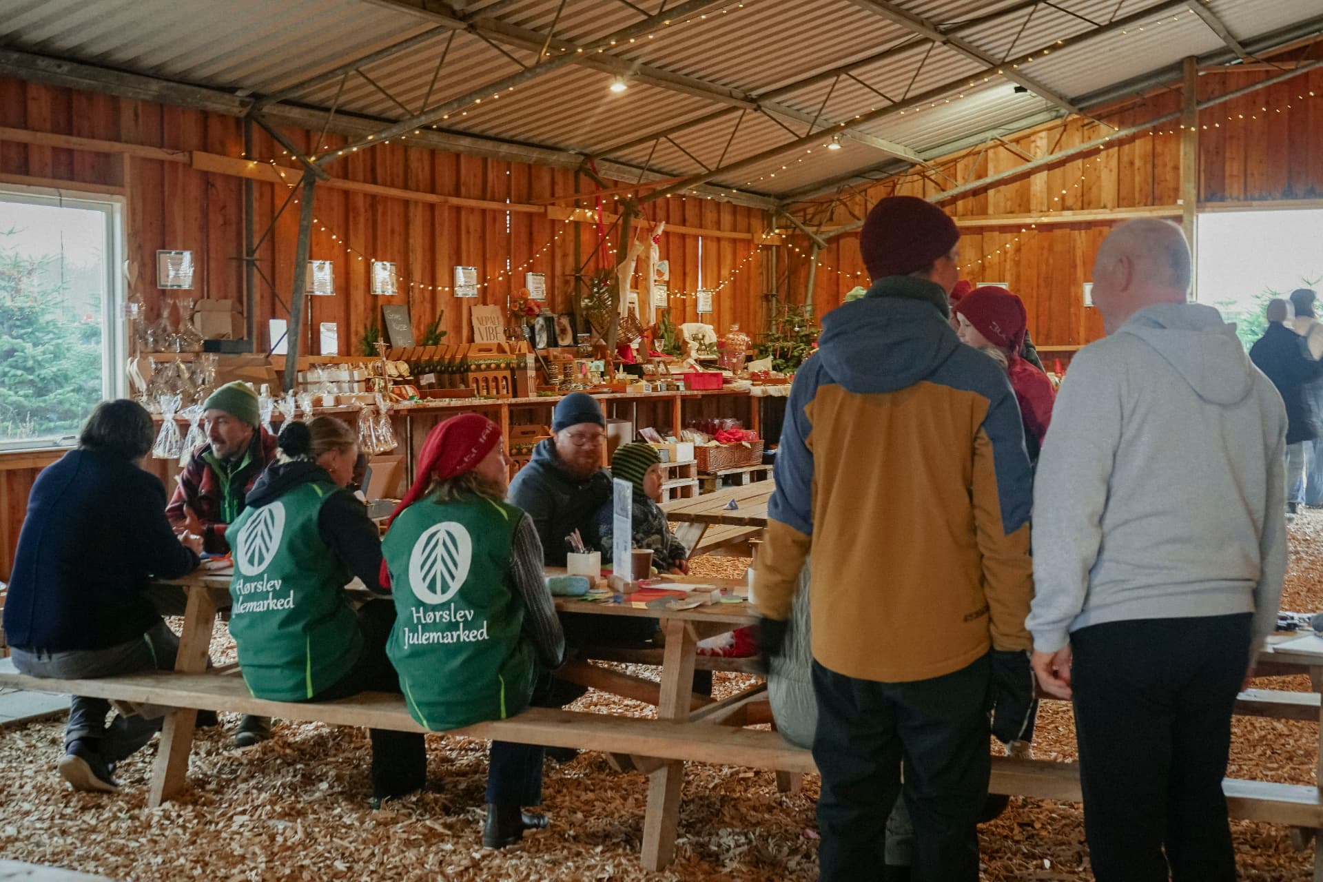 Gæster og frivillige i grønne veste ved langbordene i Træladen